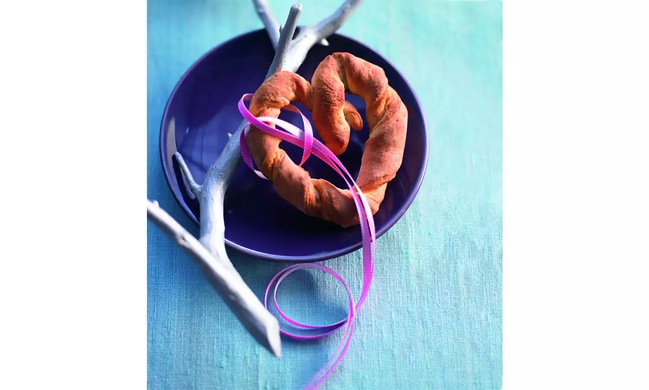 Inimioare din aluat rumen, presărate cu scorțișoară, în formă spiralată, aranjate pe o farfurie violet cu o panglică roz și crenguțe albe