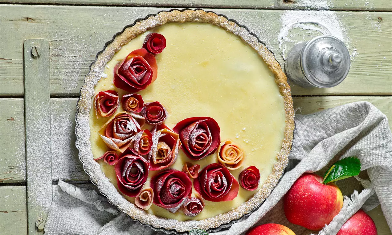 Koláč se světle žlutou jemnou náplní, okraj zlatavého těsta, zdobený růžičkami z červených jablečných plátků na rustikálním dřevěném stole