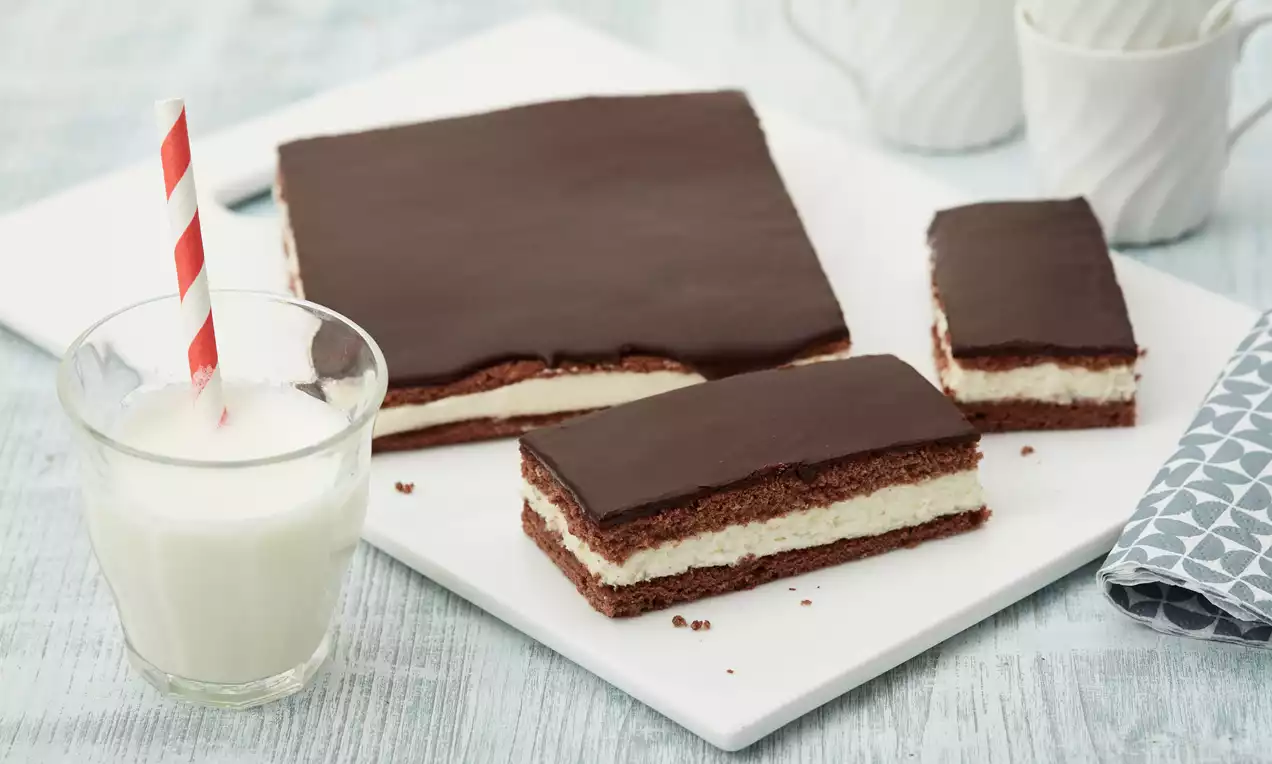 Rechteckiger Kuchen mit dunkler Schokoglasur, heller Sahnecremeschicht und lockeren Schokoladenböden auf weißer Platte mit Glas Milch