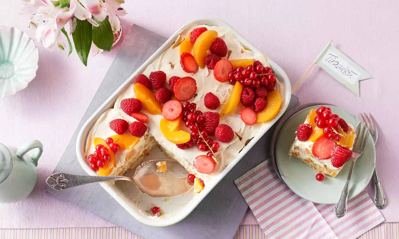 Sommerliches Tiramisu mit heller Creme, frischen Früchten wie Himbeeren, Erdbeeren und Aprikosen in einer weißen Auflaufform und auf einem Teller drapiert