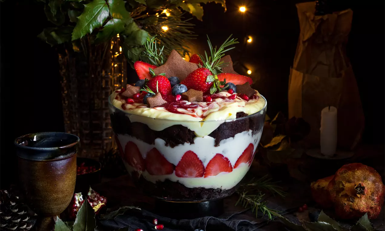 Пластове от крем светложълт и шоколадов кекс, украсени с яркочервени ягоди, боровинки и листа розмарин в стъклена купа