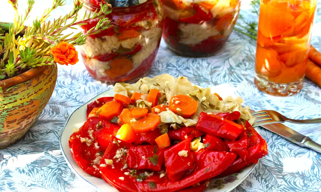 Червени парени чушки с лек блясък, гарнирани с нарязано бяло зеле и кръгчета оранжев морков върху бяла чиния с шарена покривка