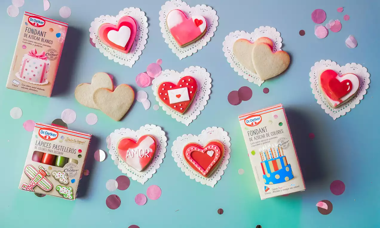 Galletas en forma de corazón decoradas con glaseado rojo, blanco y detalles románticos sobre blondas de papel y fondo azul con confeti