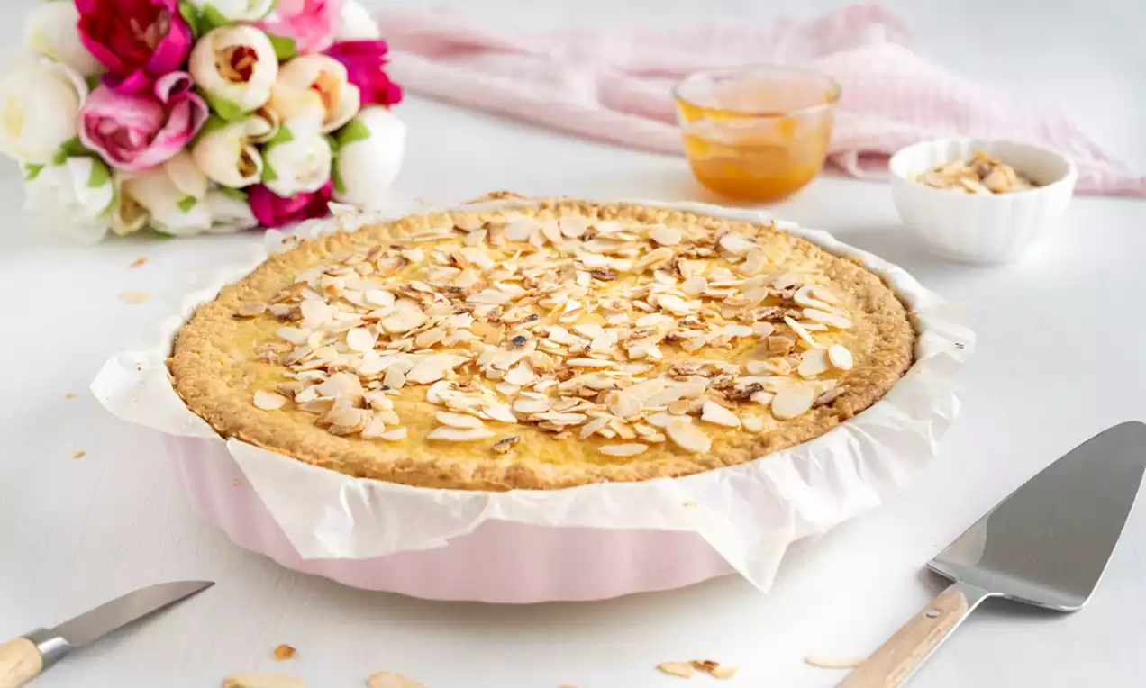 Gedeckte Vanille Pudding Tarte mit goldbrauner Oberfläche und gerösteten Mandelblättchen in einer rosa Form, daneben Blumen und Beilagen