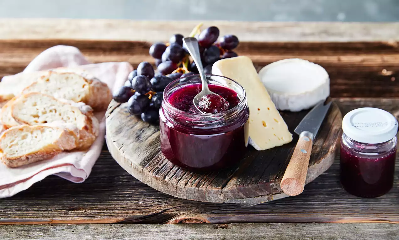 Glanzende donkerpaarse druivenconfituur in een glazen potje met lepel, gepresenteerd op een houten plank met kaas, stokbrood en druiventros