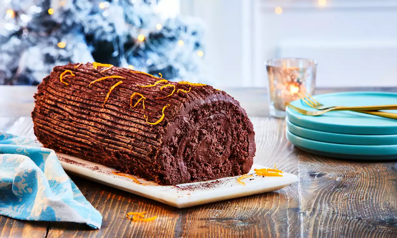 Rich chocolate vegan Yule log with a textured swirl pattern, garnished with orange zest on a white platter on a wooden table
