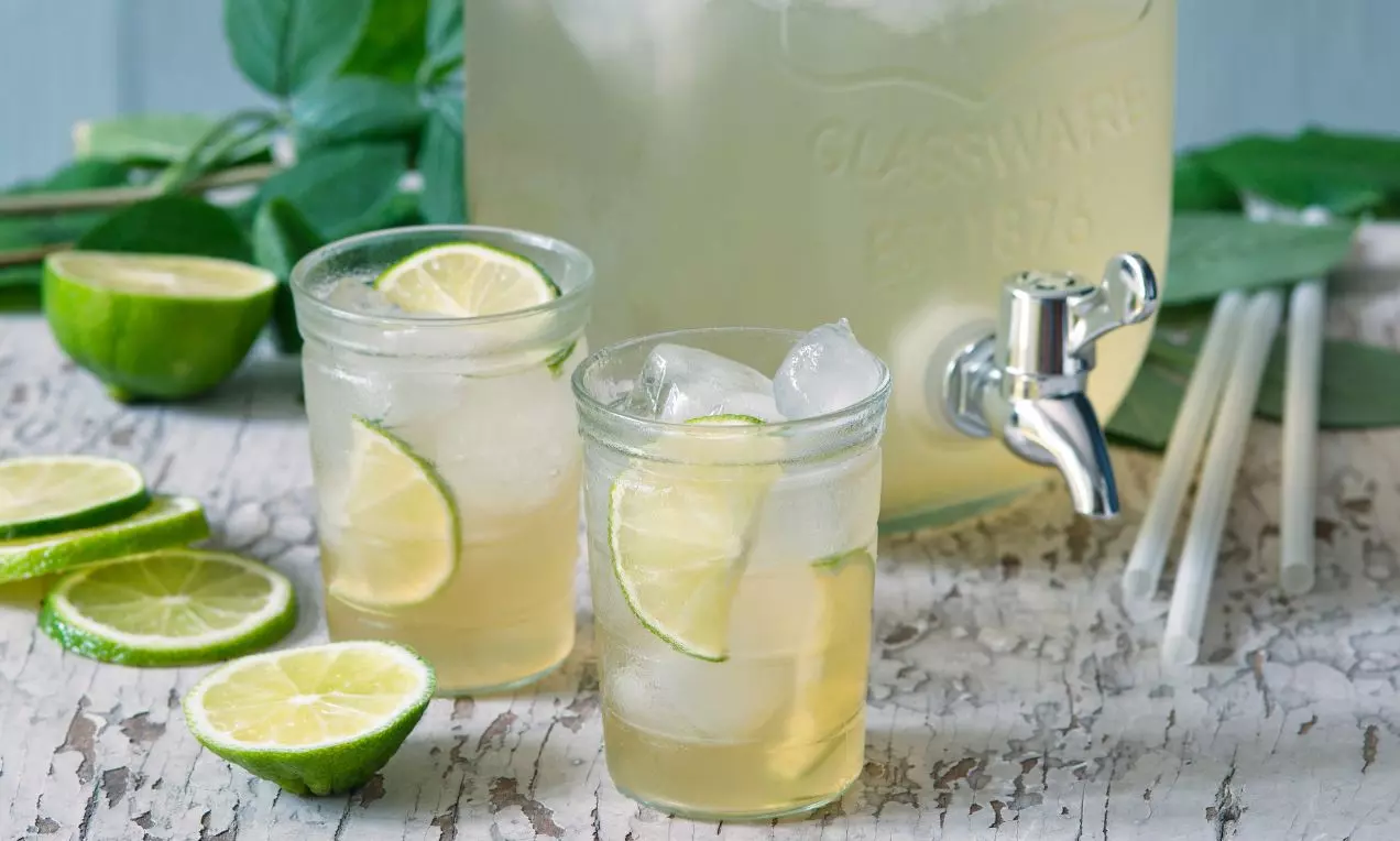 Svetložltý pomelový sirup v sklenených pohároch s ľadom a plátkami limetky na starožitnom drevenom stole vedľa zelených listov