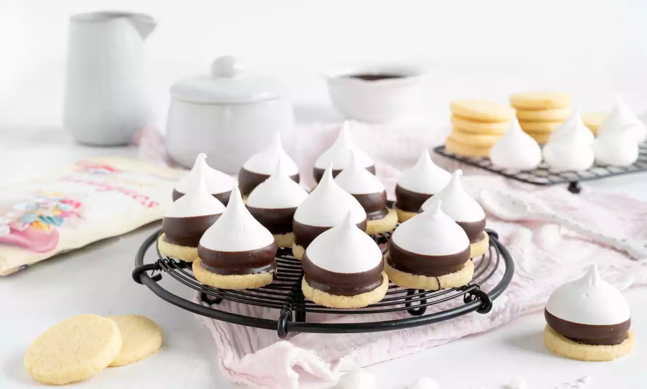 Meringue-Schoko-Cookies mit weißer glänzender Spitze, dunkler Schokoschicht und hellgelbem Keksboden auf einem schwarzen Gitter