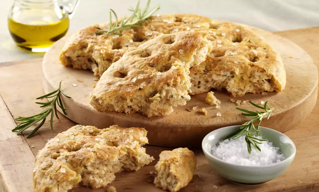 Golden vegan rosemary focaccia with a dimpled crust, broken pieces, coarse salt, fresh rosemary, olive oil and a bowl of salt on wood
