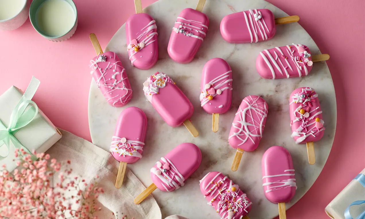 Pinke Cakesicles mit glatter Glasur, verziert mit weißen Mustern und bunten Streuseln, auf marmornem Servierteller angerichtet