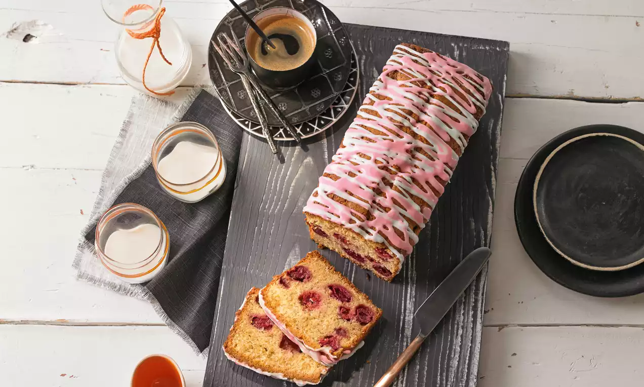 Rechteckiger Rührkuchen mit rosafarbenem Zuckerguss und weißer Glasur, durchzogen von Kirschstücken, angeschnitten auf einem grauen Holzbrett