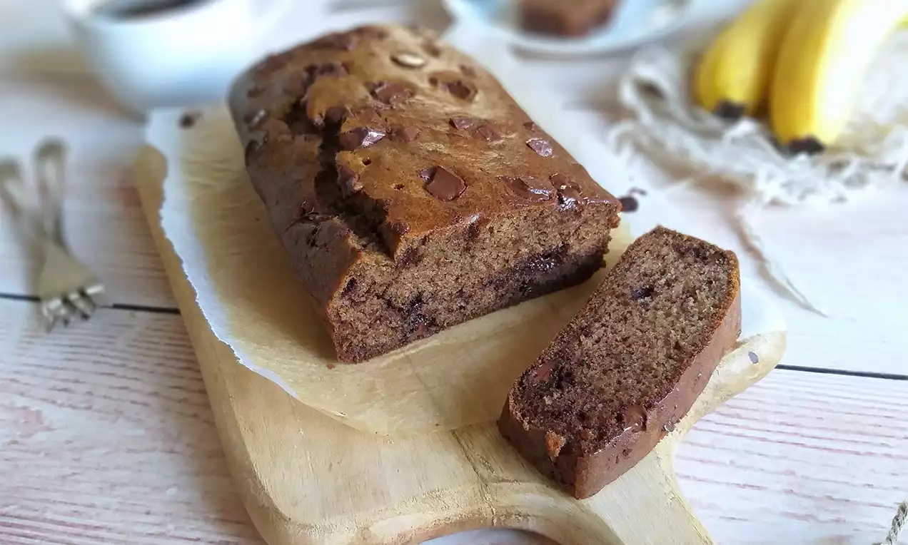 Banánový chlebíček s hnědo-zlatavou kůrkou, jemně prorostlý kousky čokolády, podélně nakrojený na dřevěném prkénku vedle zralých banánů