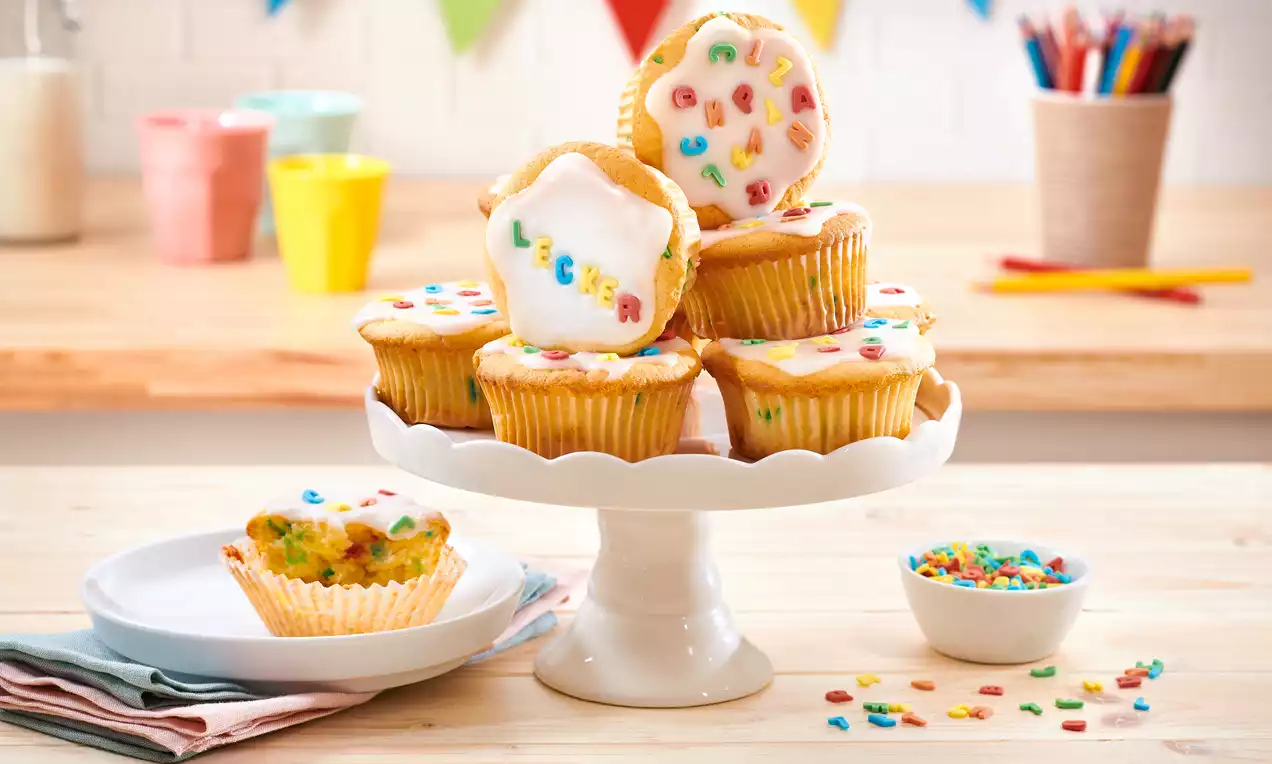 Goldgelbe Buttermilch-Muffins mit weißer Zuckerglasur und bunten Streuseln, gestapelt auf einer weißen Tortenplatte in heller Küche