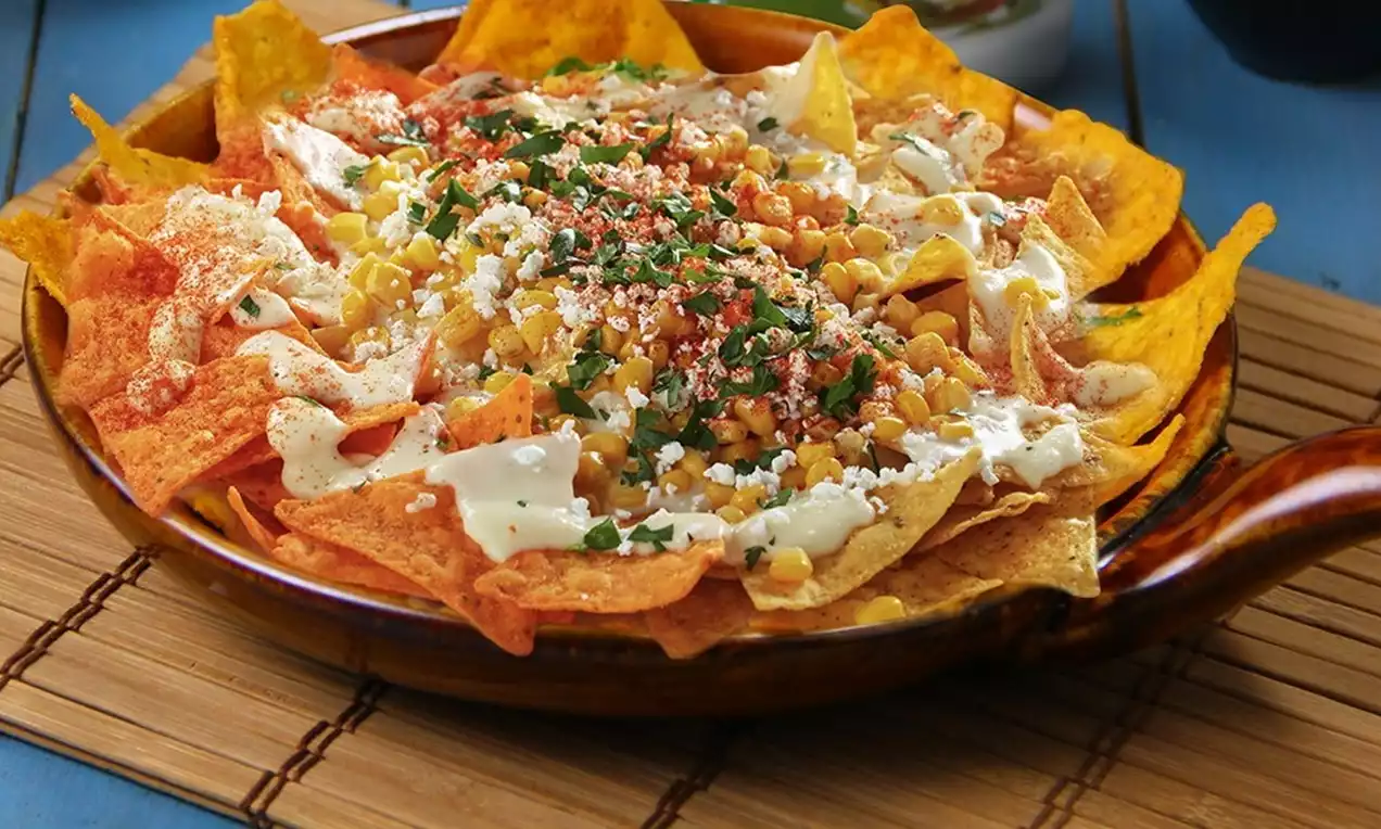 A warm, earthy-toned ceramic dish filled with crisp orange and yellow tortilla chips, layered with melted cheese, creamy sauce, golden corn, crumbled cheese, fresh green herbs and a sprinkle of red seasoning
