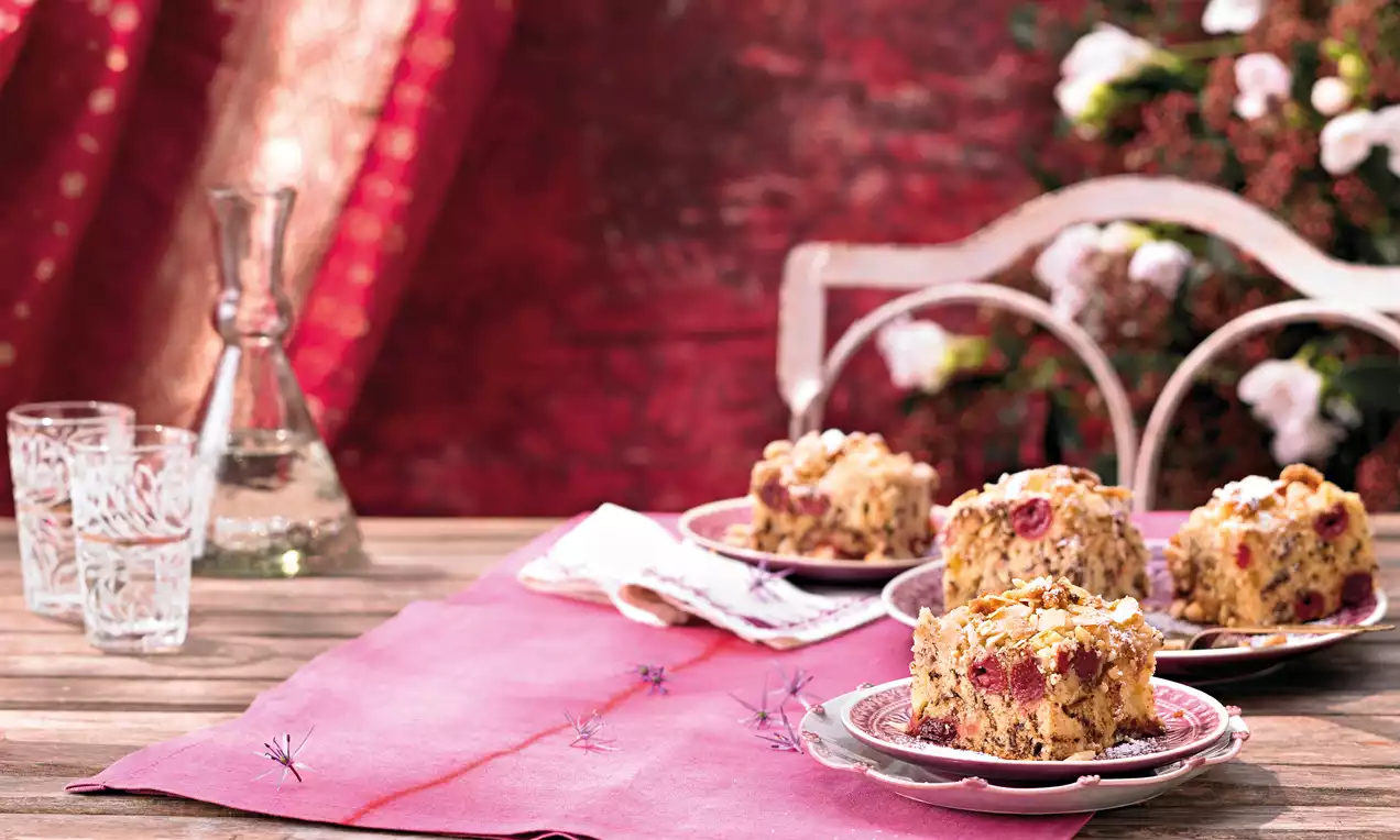 Prăjitură cu vișine suculente pe aluat sfărâmicios auriu, presărată cu fulgi de migdale, servită pe farfurii decorative roz pe o masă rustică