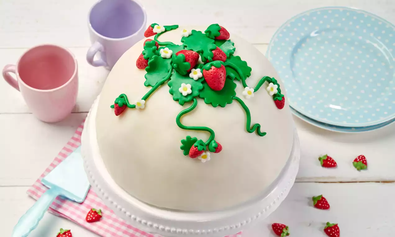 Runde Kuppeltorte mit weißem Überzug, verziert mit grünen Blättern, roten Erdbeeren und kleinen weißen Blüten, auf weißer Tortenplatte