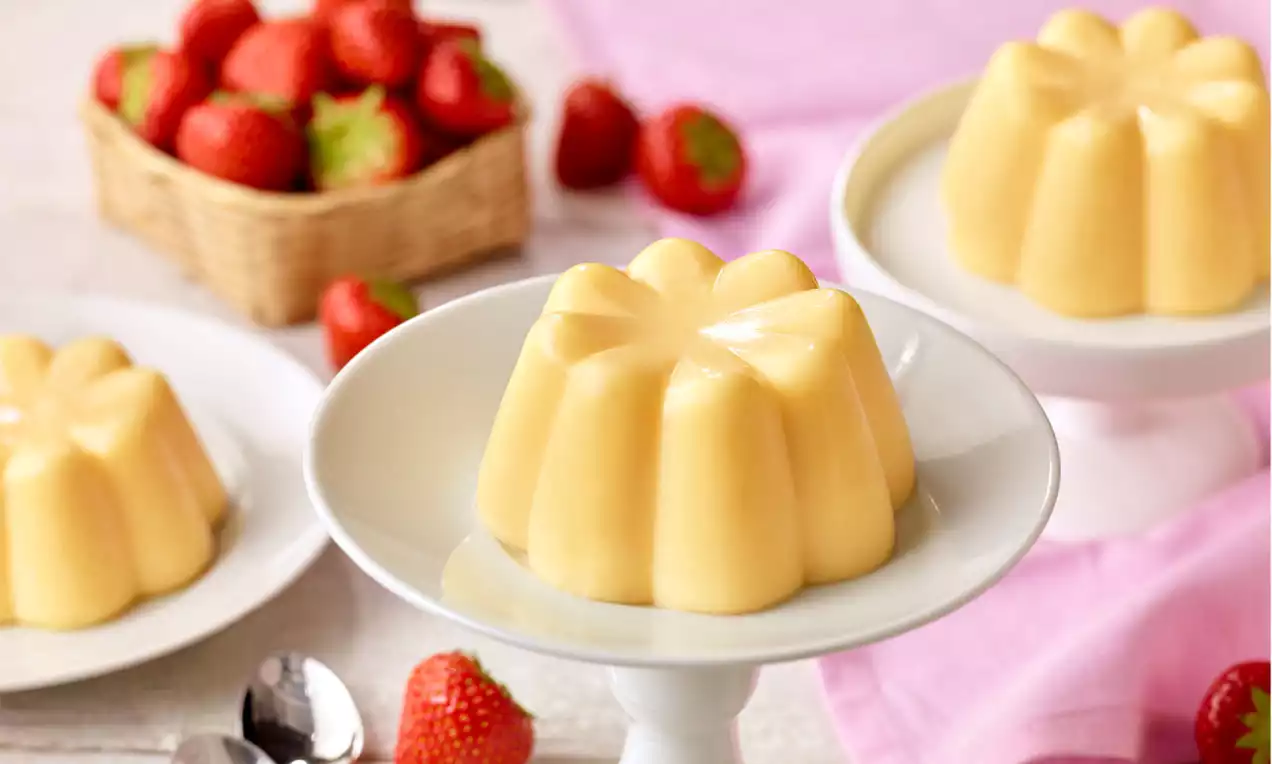 Gelber Vanillepudding in Blumenform auf einer weißen Dessertschale, umgeben von roten Erdbeeren und rosa Stoff auf hellem Hintergrund