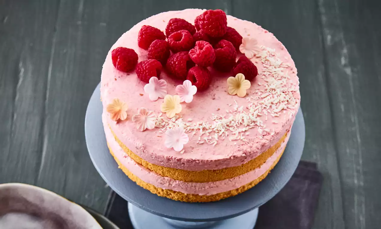 Himbeer-Joghurt-Torte mit rosafarbener Creme und gelbem Biskuitboden, dekoriert mit frischen Himbeeren, Kokosraspeln und Blüten auf einer blauen Tortenplatte