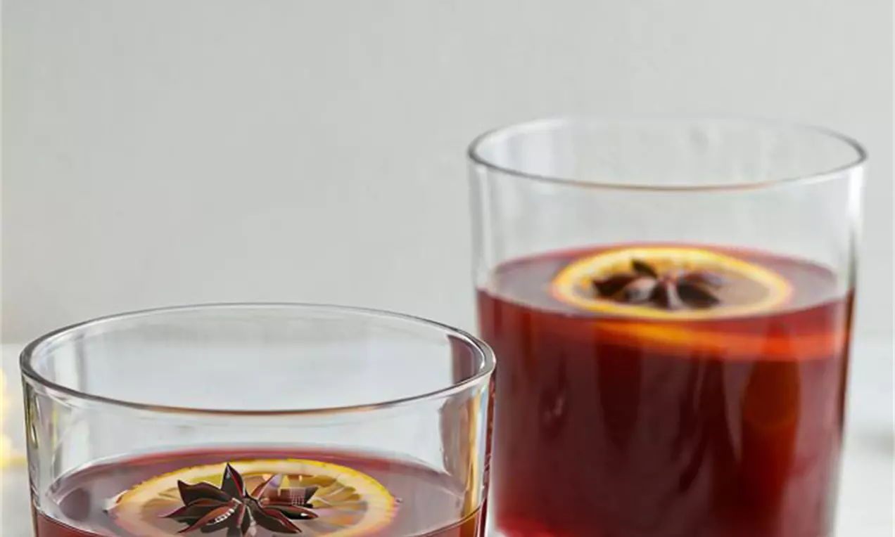 Picture - Clear glasses filled with deep red vanilla glögg, each garnished with a slice of lemon and a star anise, surrounded by spices on a white surface