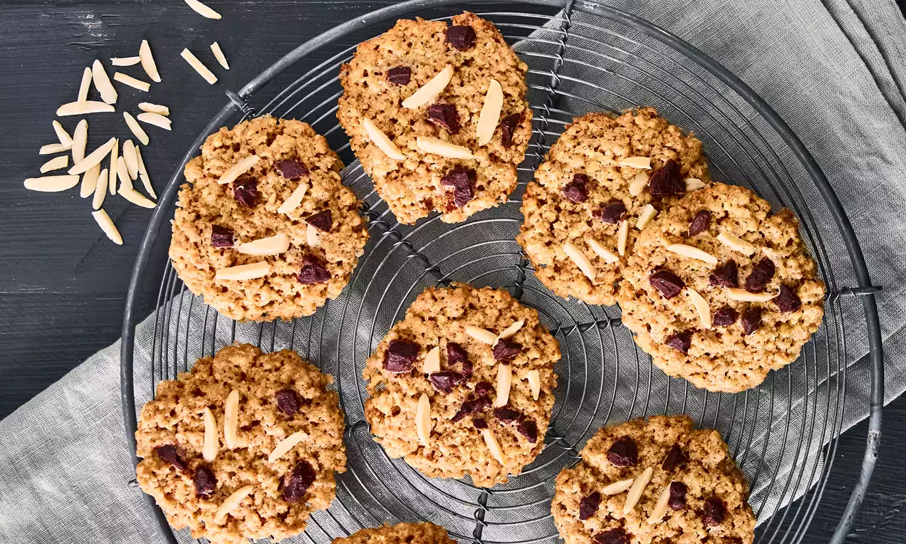 Runde, goldbraune Schoggi-Dinkel-Cookies mit Stücken aus dunkler Schokolade und gehobelten Mandeln auf einem schwarzen Gitterrost