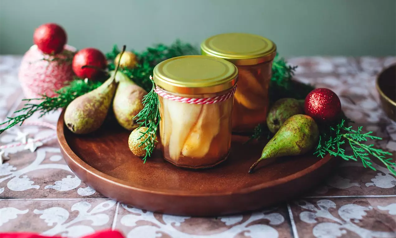 Zlatistý hruškový kompót v sklenených pohároch so zlatými viečkami, obklopený čerstvými hruškami, vetvičkami ihličia a červenými ozdobnými guľami