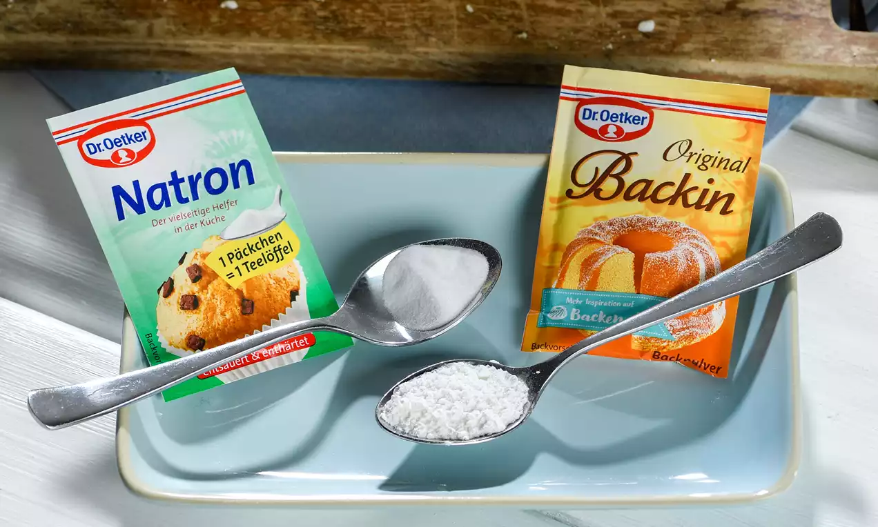 Zwei silberne Löffel mit weißem Natron und Backpulver auf hellblauer Keramikplatte, daneben die passenden Verpackungen mit farbigen Motiven
