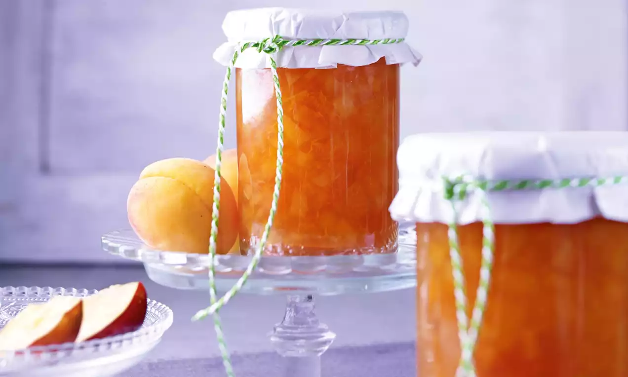 Heldere potten met oranje abrikozenjam, wit papierdeksel met groen touw, op een glazen taartstandaard met abrikozen en gesneden plakjes