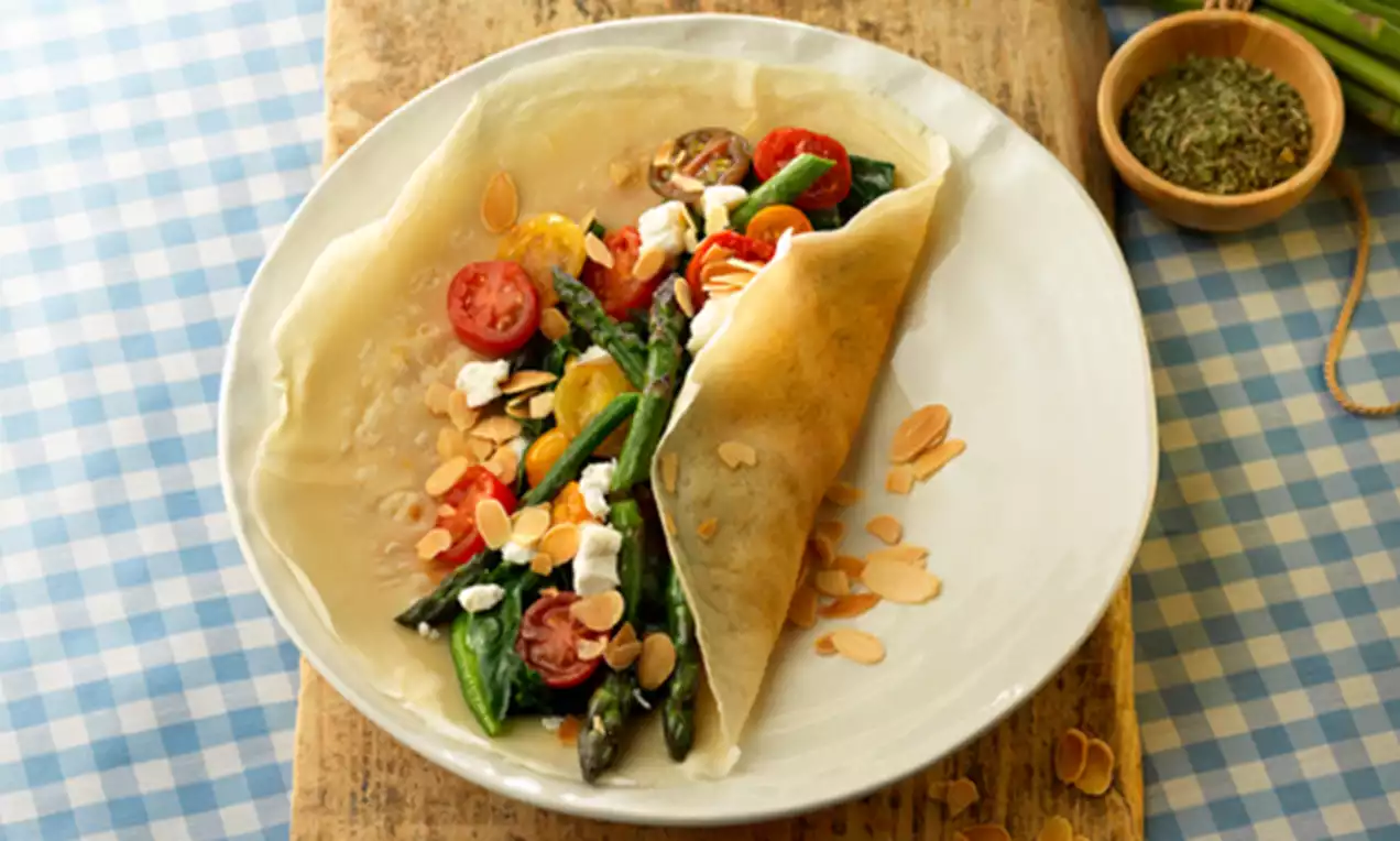 Dunne goudbruine crêpe gevuld met kleurrijke spinazie, tomaat, asperges, fetakruimels en amandelschaafsel op een wit bord op een houten plank
