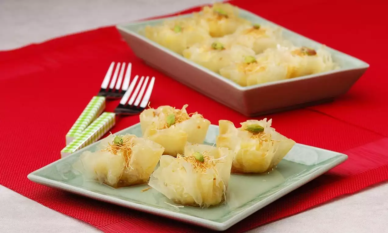 Kare şekilli nane yeşili tabakta sunulan altın sarısı çıtır baklavalar, üzerileri antep fıstığıyla süslenmiş ve arka planda kırmızı örtü bulunuyor