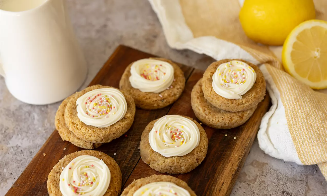 Citroencookies met ronde, goudbruine basis en witte roomtopping met gekleurde sprinkles op een houten plank naast citroenen en linnen doek