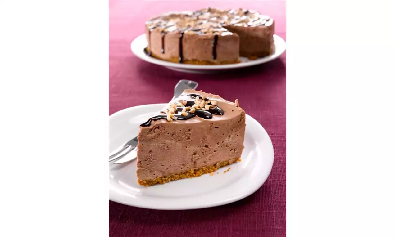 Sjokoladekake med nøttebiter på gyllen kjekssbunn, toppet med mørk sjokoladesaus og ristede nøtter på hvitt fat mot rosa bordduk