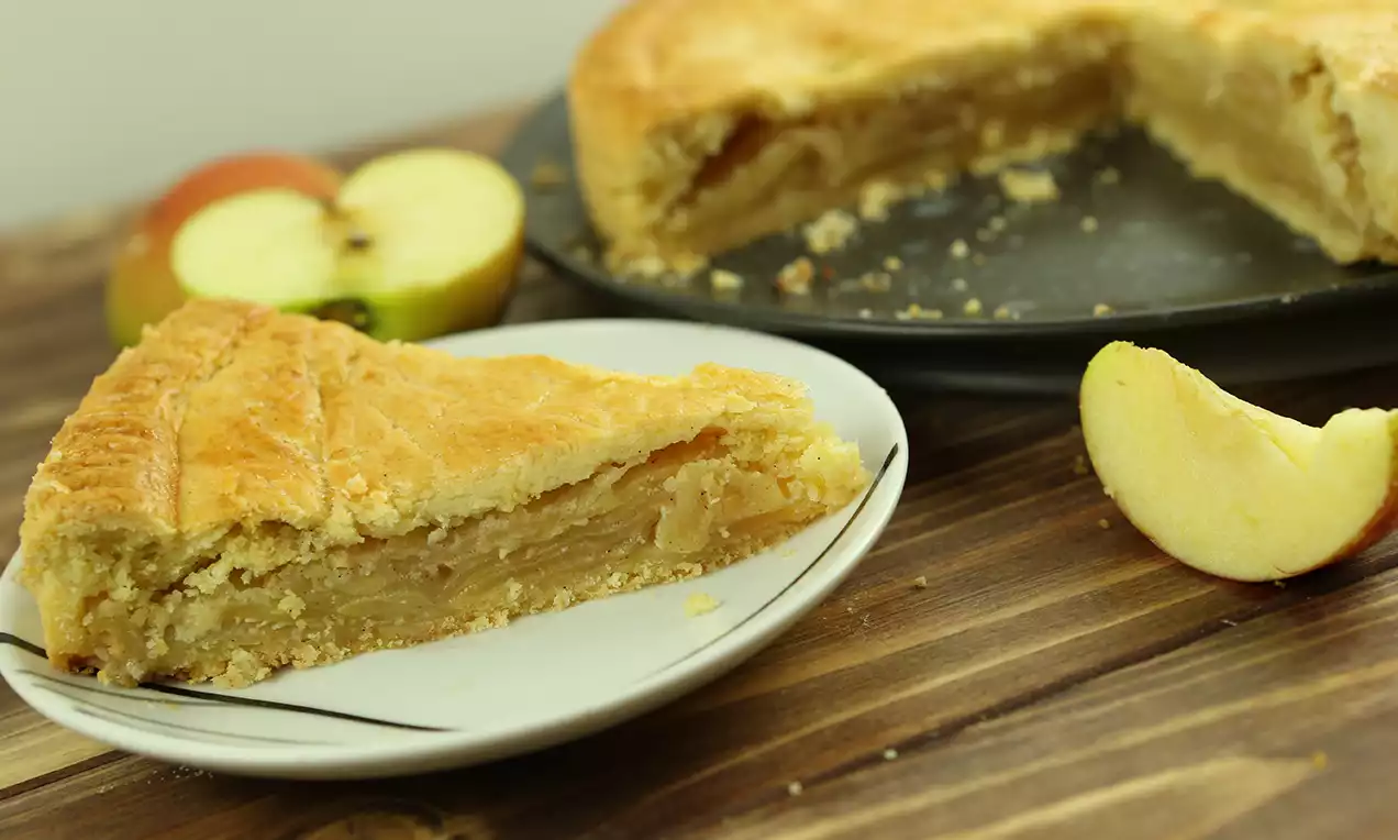 Ein goldbrauner Kuchenschnitt mit zart-blättriger Kruste und fruchtiger Apfelfüllung auf weißem Teller neben frischen Apfelscheiben