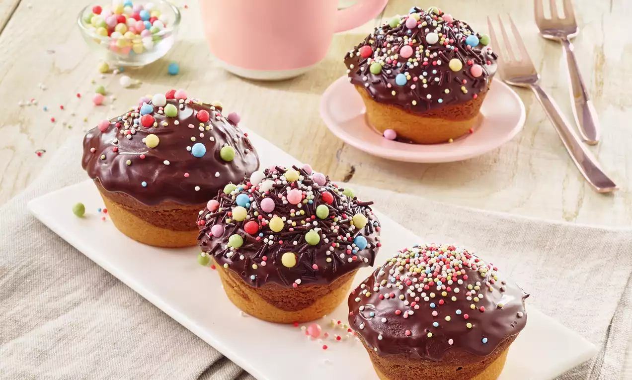 Muffini sa smeđom čokoladnom glazurom, ukrašeni šarenim perlicama, posluženi na bijelom tanjuru s dvije šalice kave u ružičastim šalicama