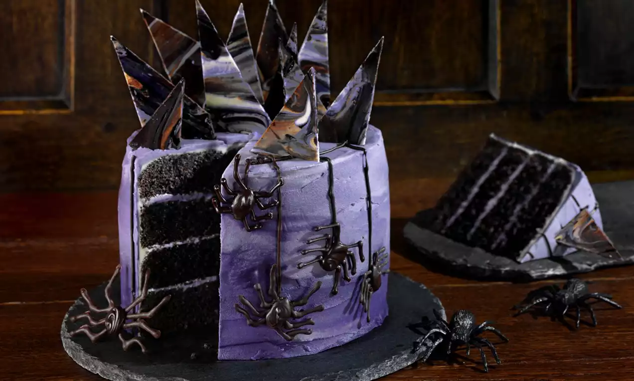 A layered cake with deep purple frosting, sharp marbled chocolate shards, black spiders, and dark sponge visible on a slate plate