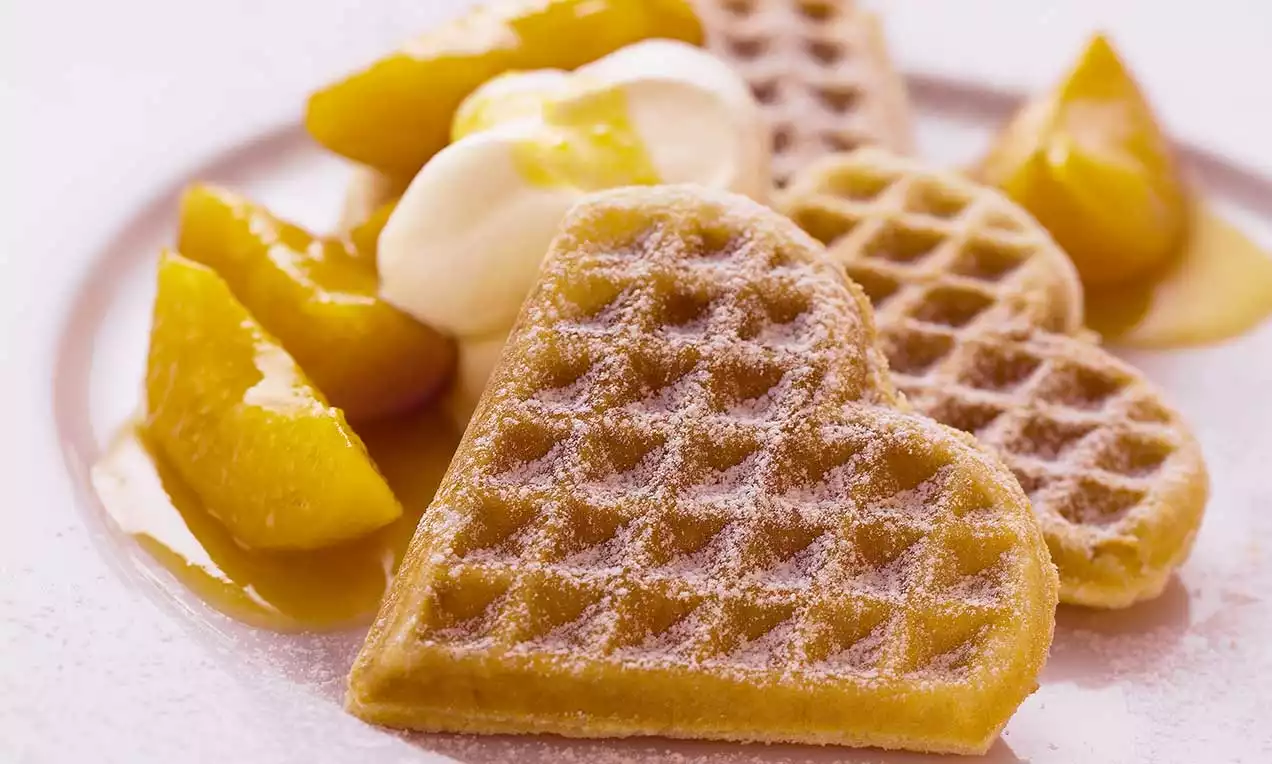 Herzförmige Hefewaffeln goldbraun gebacken, mit Puderzucker bestreut, neben Sahne und glänzenden, gelben Pfirsichspalten angerichtet