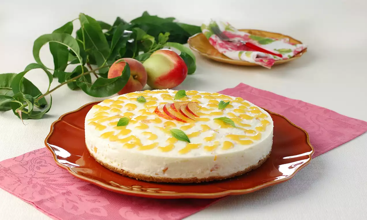 Yuvarlak kahverengi tabakta, beyaz kremalı, altın şeftali dilimleri ve nane yapraklarıyla süslenmiş, bisküvi tabanlı cheesecake