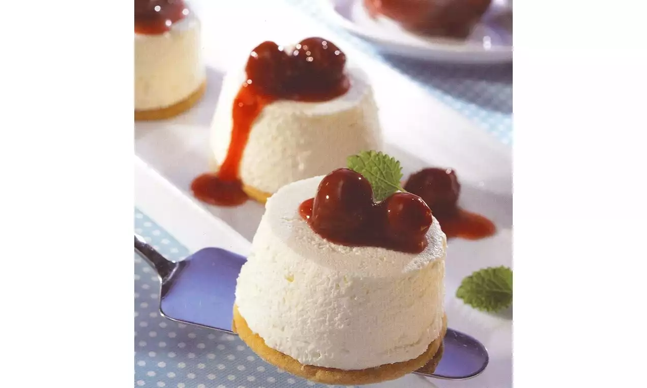 Petites timbales de mousse blanche sur un socle biscuité, garnies de cerises rouge vif et de coulis brillant, décorées de feuilles de menthe