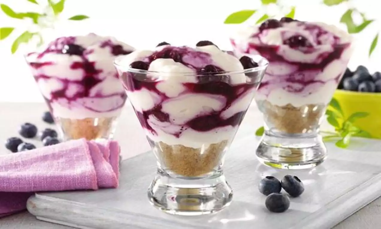 Dessert im Glas mit Schichten aus hellem Keksboden, weißer Creme und dunkelvioletter Blaubeersauce, dekoriert mit frischen Blaubeeren