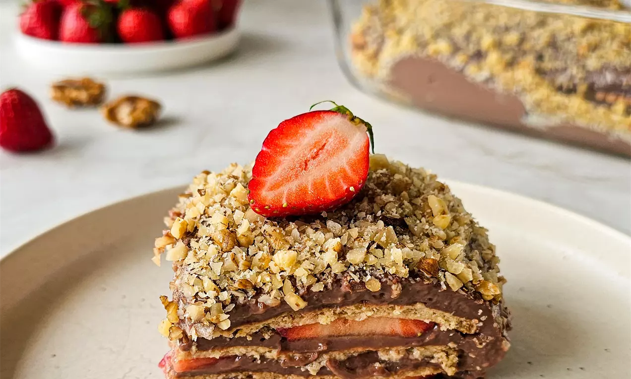 Слоеста торта с бисквити, червени резени ягоди и шоколадов крем, покрита с хрупкави ядки, поднесена върху светла чиния