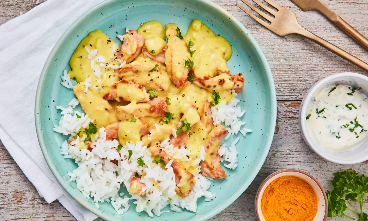 Putengeschnetzeltes in cremiger gelber Sauce mit heller Textur auf weißem Reis, angerichtet in einer türkisfarbenen Schale mit Petersilie garniert