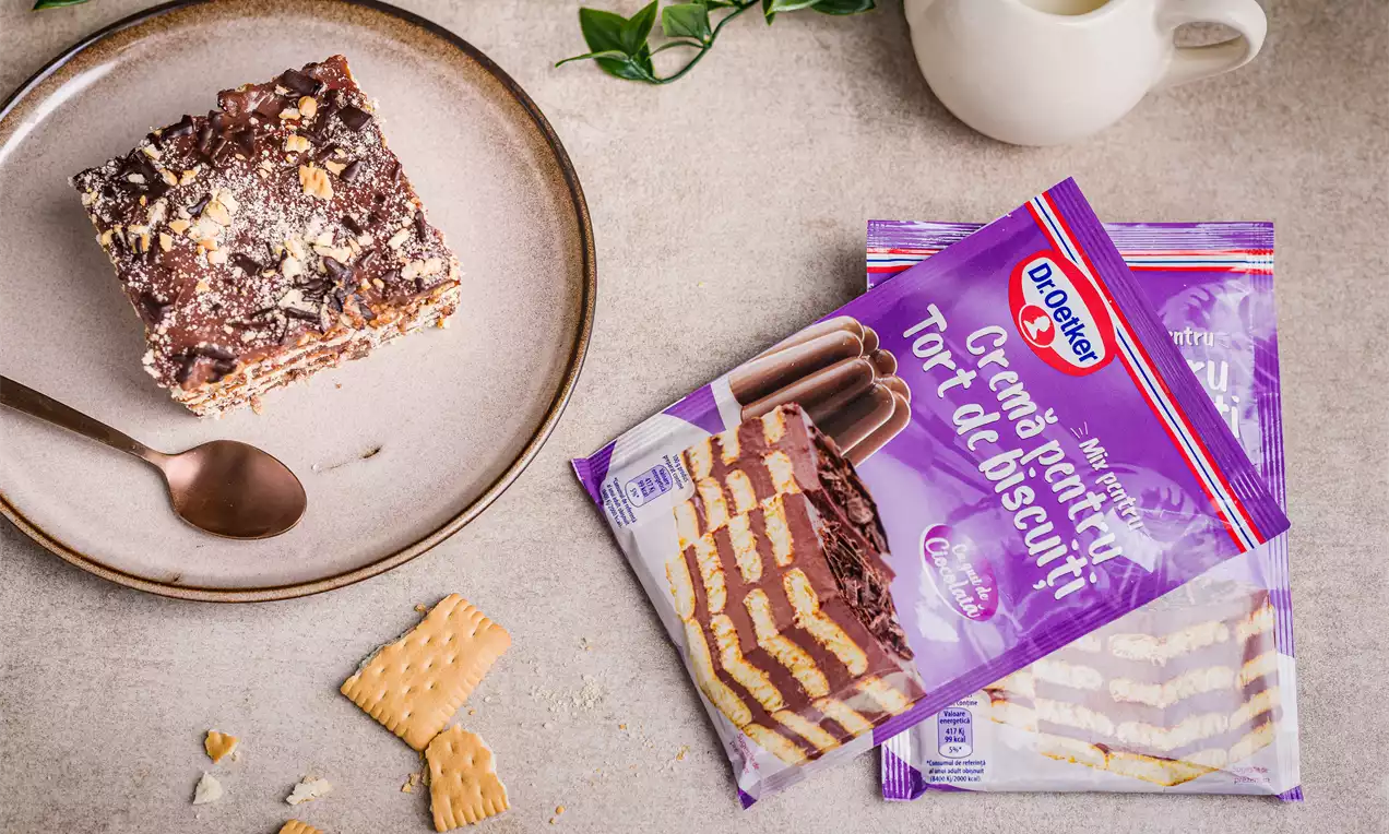 Tort dreptunghiular cu straturi alternante de biscuiți aurii și cremă de ciocolată maro închis, decorat cu nuci mărunțite pe o farfurie bej