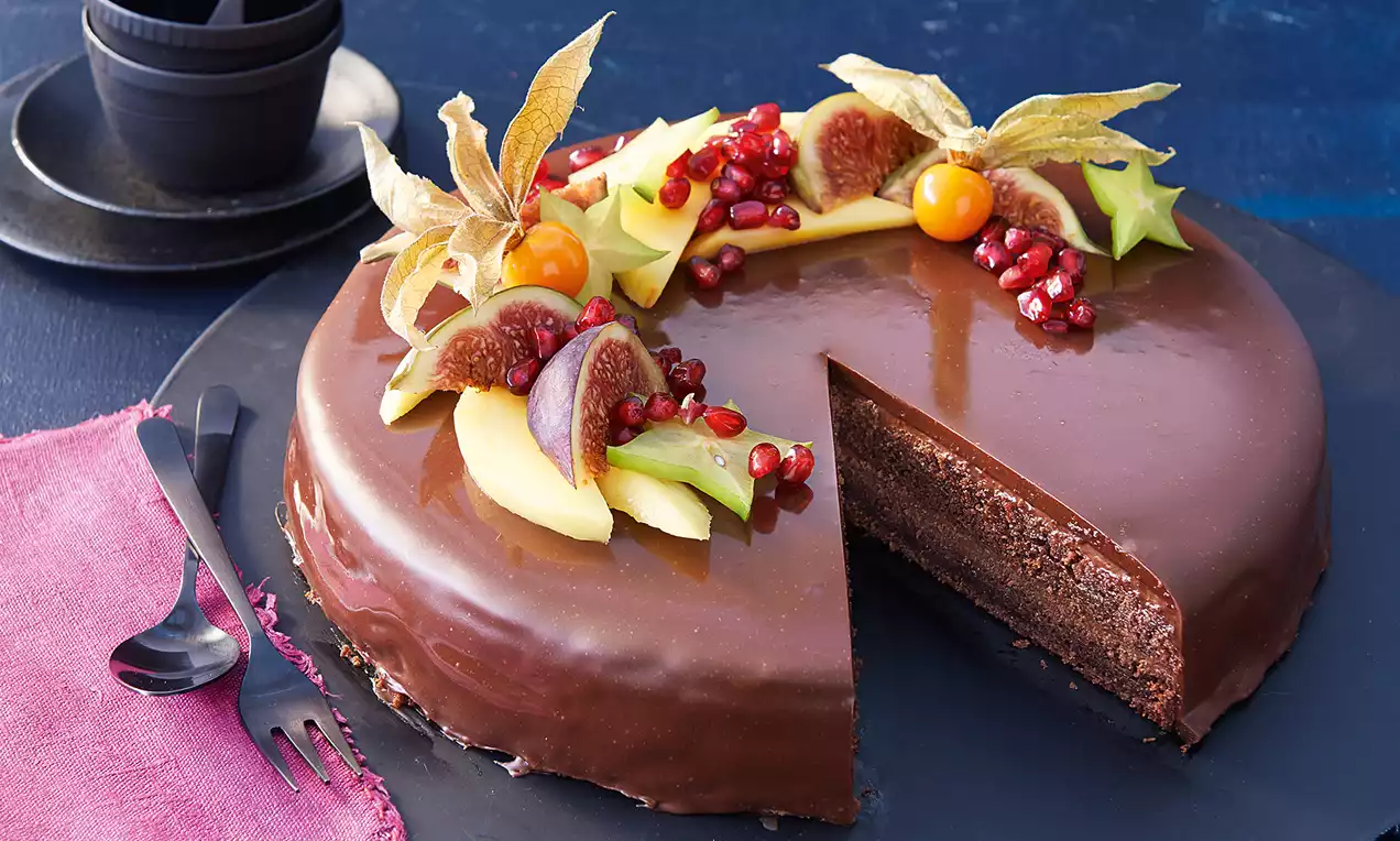 Schokoladentorte mit glatter, glänzender Oberfläche, dekoriert mit Obst wie Physalis, Feigen, Granatapfelkernen und Sternfrucht auf schwarzer Platte