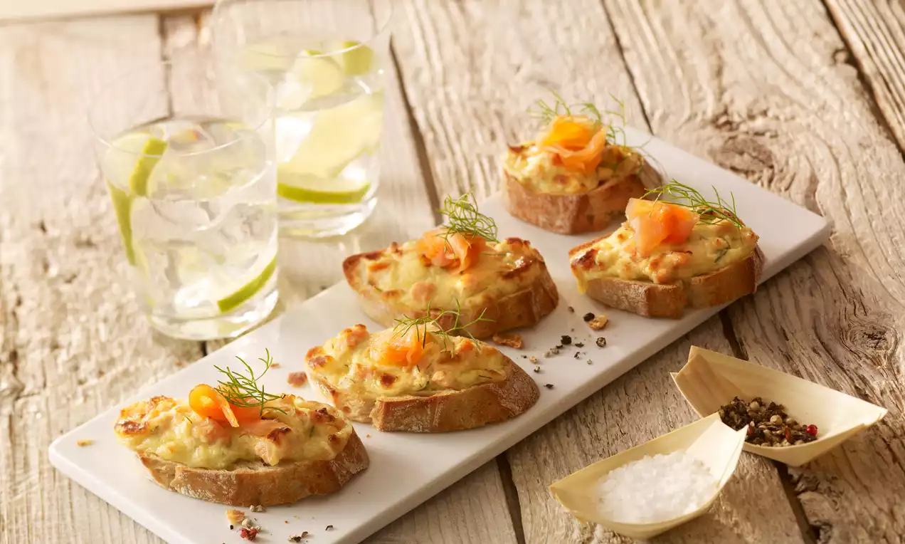 Überbackenes Lachs-Baguette mit rosa Lachs, goldbraunem Käse, cremiger Senf-Joghurt-Honig-Creme, Dill und Zitrone auf rustikalem Holzbrett