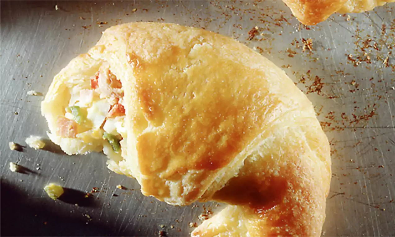 Goudbruin gebakken croissant in een halve cirkel met een vulling van roze ham en groene stukjes groenten op een metalen bakplaat