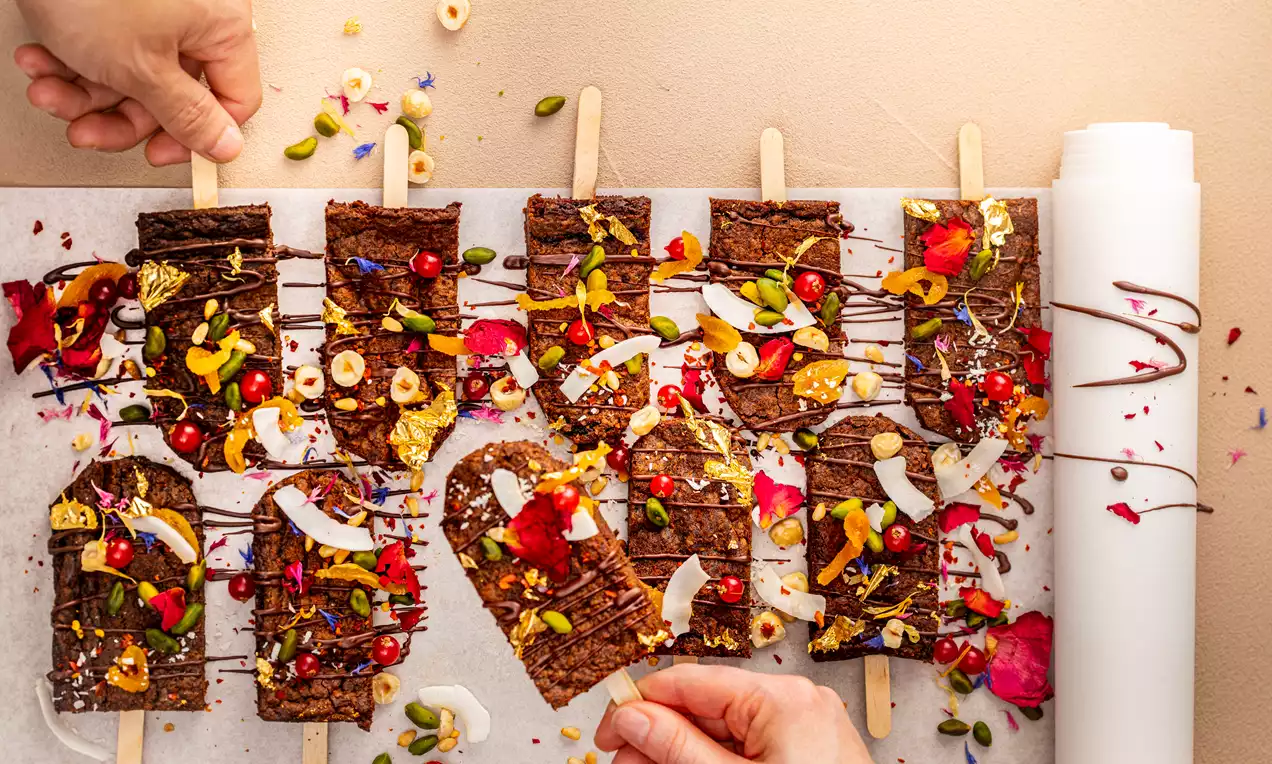 Sucettes de brownies carrés à la texture chocolatée, garnis de pétales colorés, éclats de fruits secs et morceaux de chocolat sur du papier cuisson