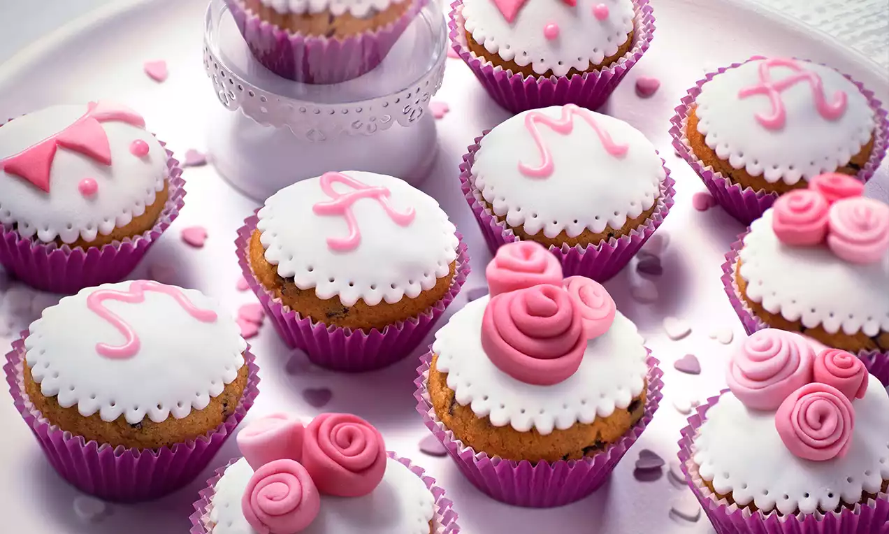 Morbidi muffin dorati in pirottini rosa, decorati con glassa bianca, dettagli rosa e forme di rose e lettere, su un vassoio chiaro