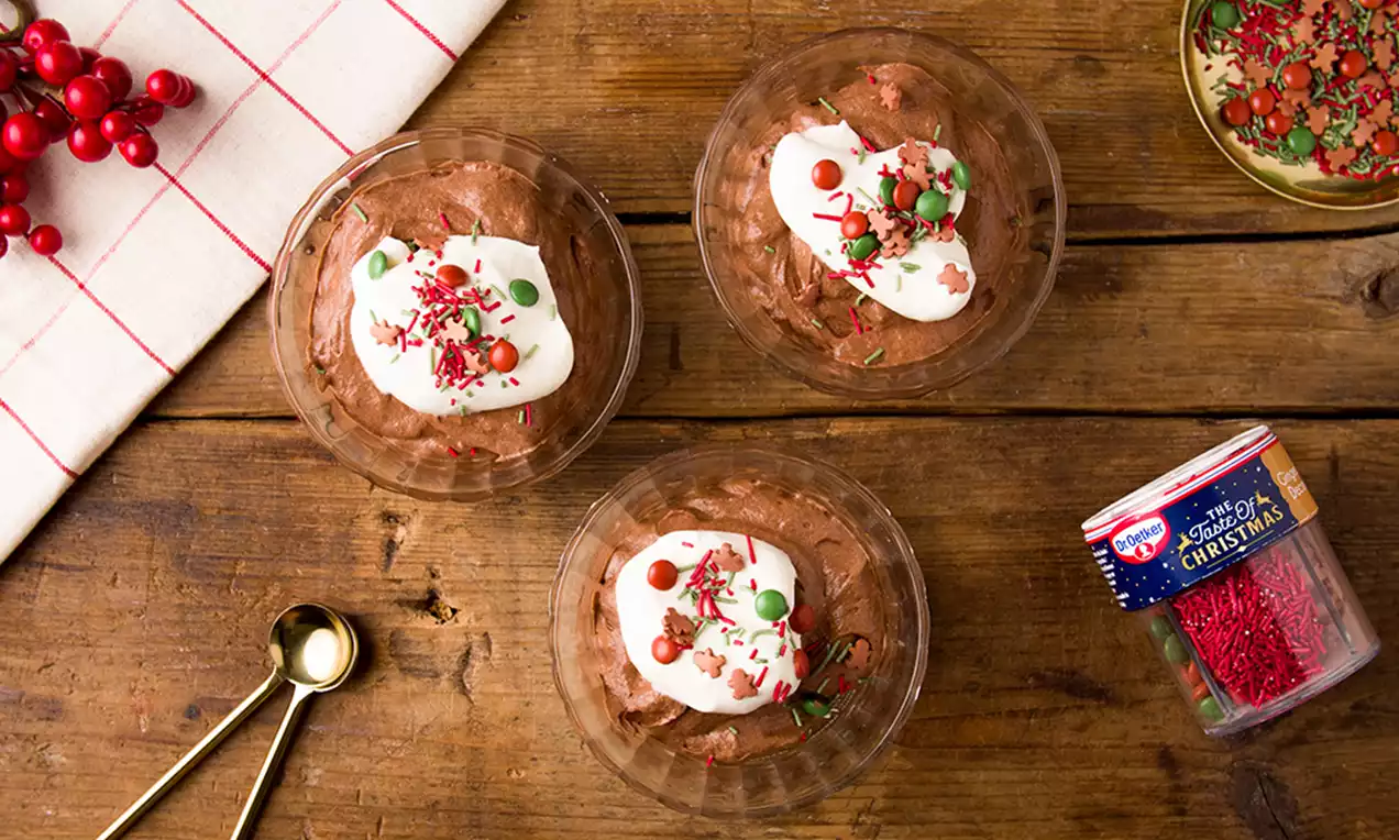 Mousses de chocolate marrón oscuro en vasos plásticos, coronados con crema blanca, chispas rojas y verdes y decoraciones navideñas