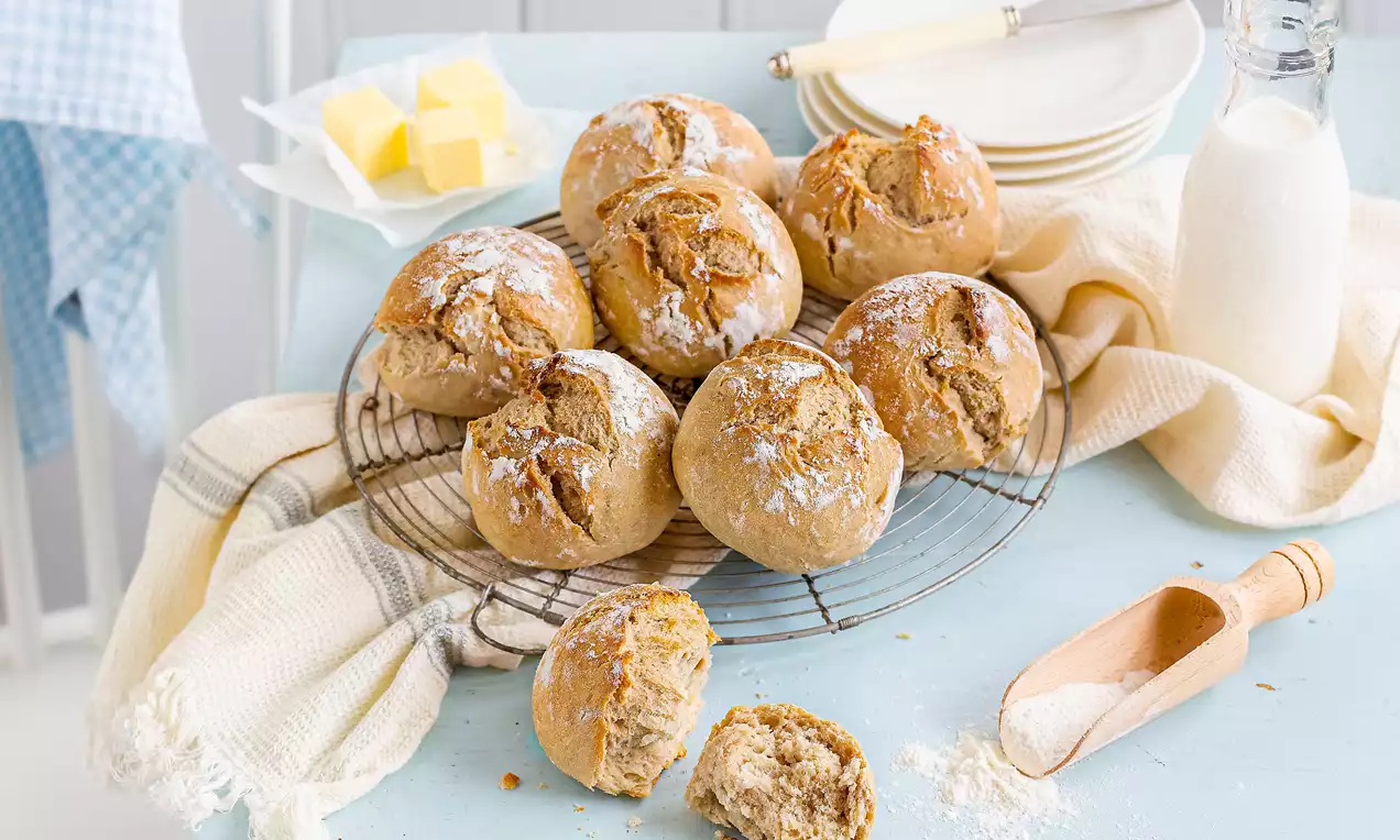 Goldbraune Sauerteig-Brötchen mit rissiger Kruste auf einem runden Rost, umgeben von Butterwürfeln, einer Milchflasche und Mehl auf hellem Untergrund