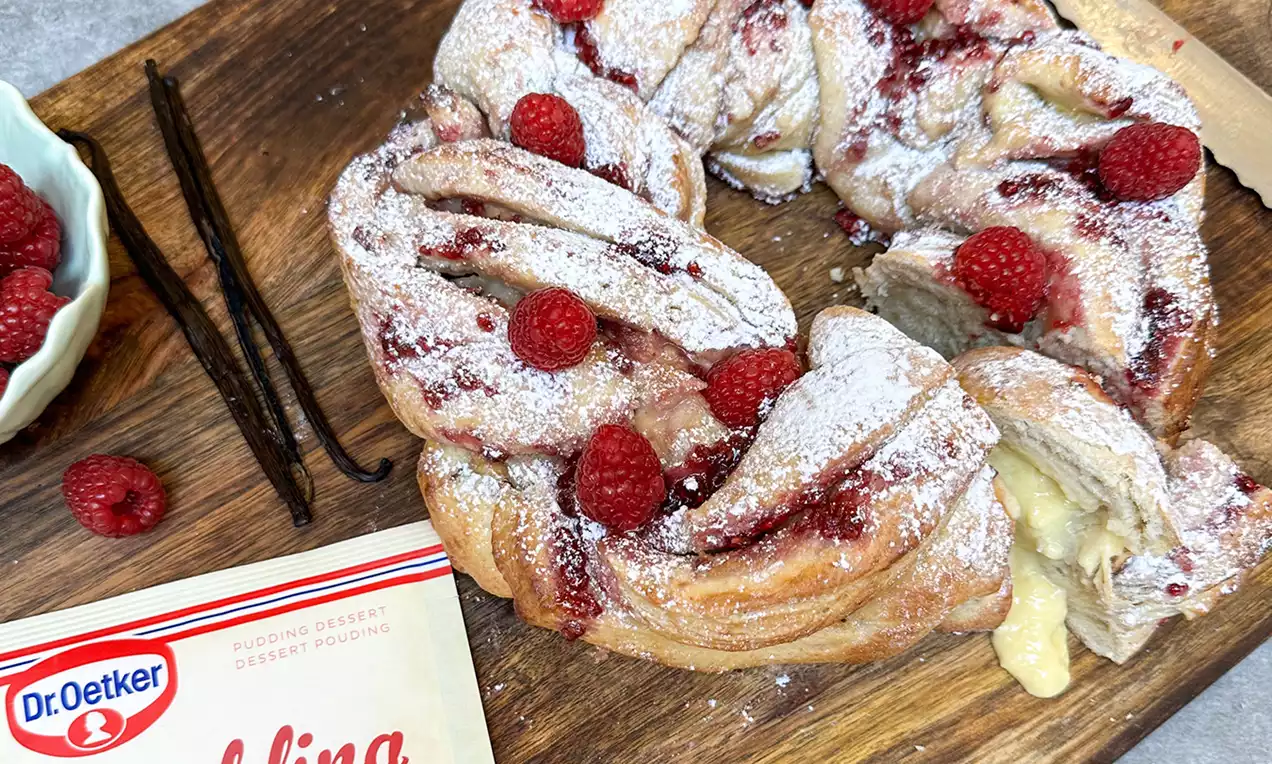 Goldbrauner, zopfartig geflochtener Vanillepudding-Kranz mit weißem Puderzucker und frischen roten Himbeeren auf einem Holzbrett drapiert