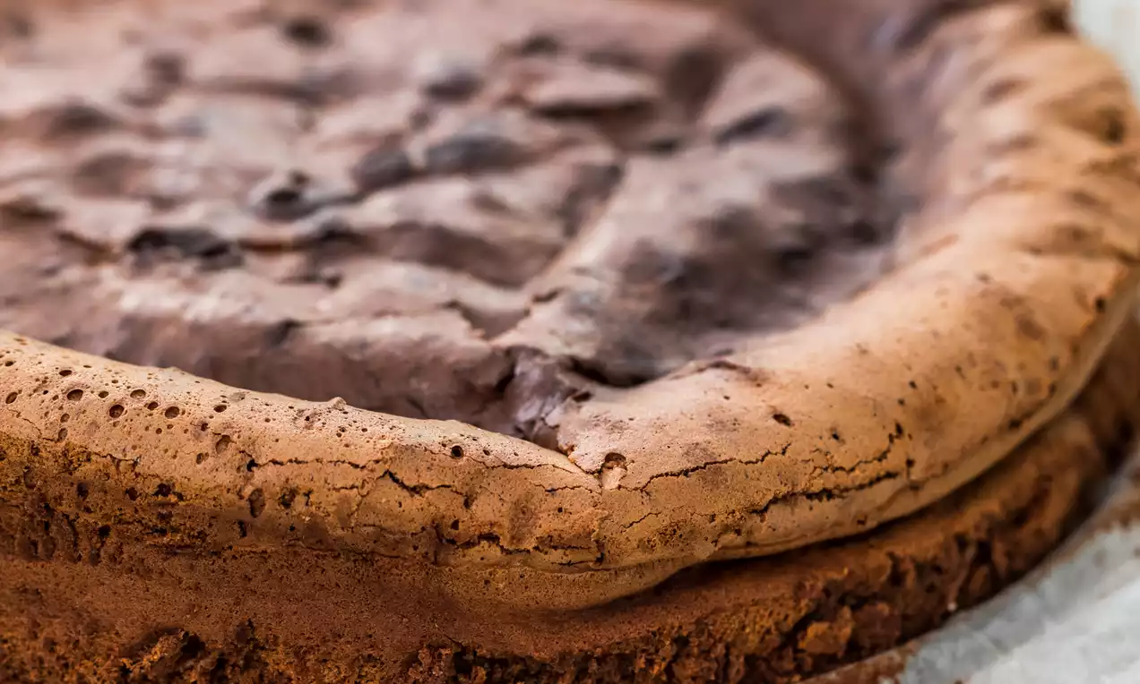 Bizcocho redondo de chocolate oscuro con textura agrietada y borde ligeramente elevado, sobre papel de horno blanco